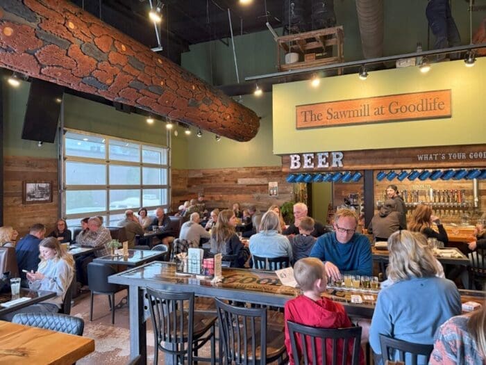 a Lively Restaurant Interior with Wooden Decor and a Faux Log Beam Diners Sit at Tables Chatting and Eating a Sign Reads the Sawmill at Goodlife Above a Bar Area Displaying Beer the Atmosphere is Warm and Social with Families and Groups Gathered