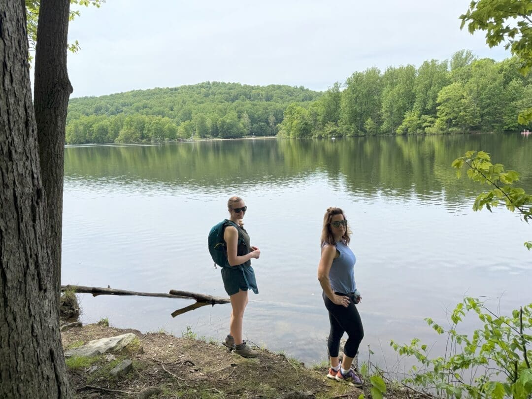 Two women stand by a calm lake at Cunningham Falls State Park, surrounded by lush green trees and hills. One wears a backpack and shorts, the other leggings and sunglasses. Both face the camera, with reflective water and forested shoreline behind them.