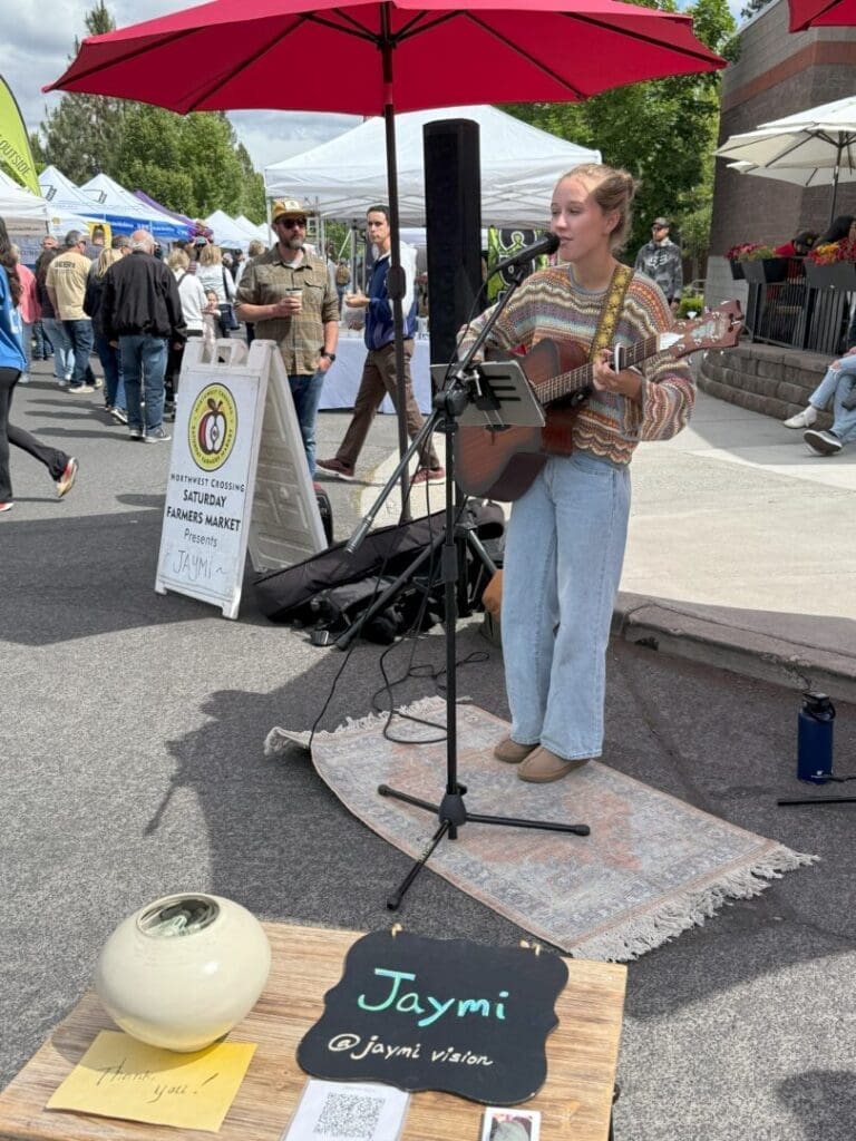 a Young Woman Stands on a Rug Playing Guitar and Singing into a Microphone at an Outdoor Market She Wears a Striped Sweater Light Jeans and Beige Shoes a Sign in Front Reads Jaymi People Tents and an Umbrella Are Visible in the Background