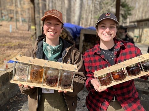 Two smiling people outdoors at Cunningham Falls State Park hold wooden trays with four jars each, filled with amber and brown liquids. They wear casual clothes, hats, and flannel shirts, with leafless trees and stacked wood in the background.
