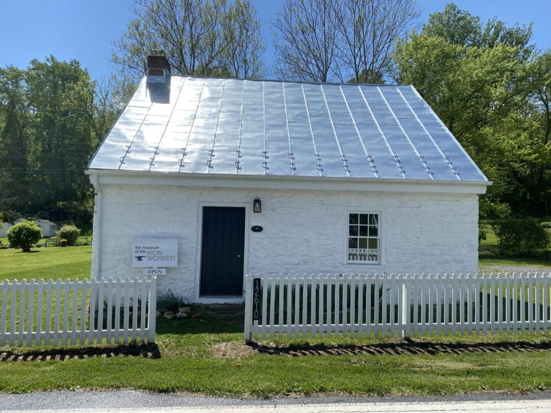 A small white stone house with a shiny metal roof, black door, and single window sits near Cunningham Falls State Park. A white picket fence surrounds the yard, with green trees and lawn under a clear blue sky.