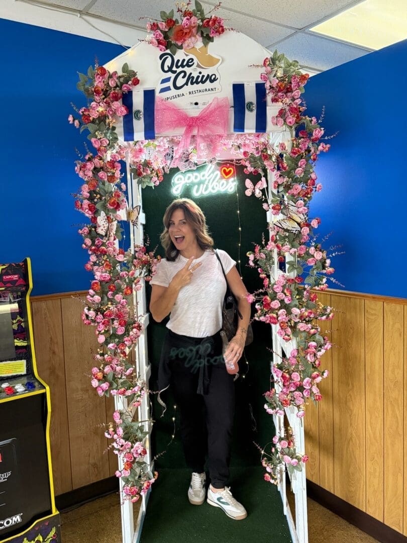 a Smiling Woman Stands Under a Floral Archway Decorated with Pink Flowers Leaves Salvadoran Flags and a Neon Good Vibes Signproudly Pointing to Herself As if to Say She Knows Where to Find the Best Pupusas in Frederick Md Wood Paneled Walls and an Arcade Machine Complete the Scene