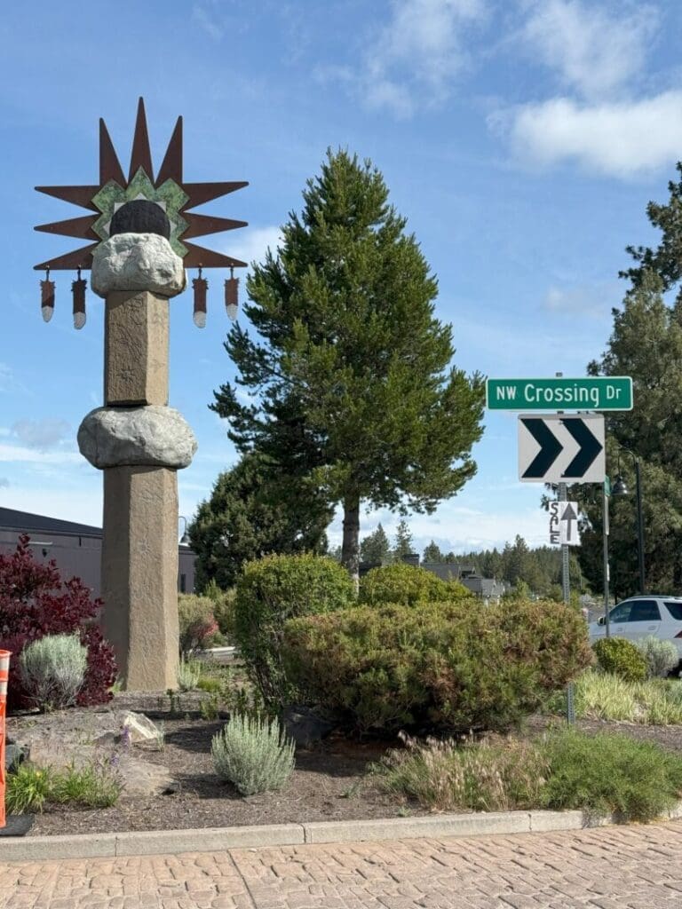 a Tall Roadside Sculpture with Two Large Rocks Stacked on a Pillar Topped by a Sunburst Design and Four Hanging Lanterns It Stands by a Street Sign Reading Nw Crossing Dr and a Right Turn Arrow Surrounded by Greenery and Trees Under a Partly Cloudy Sky