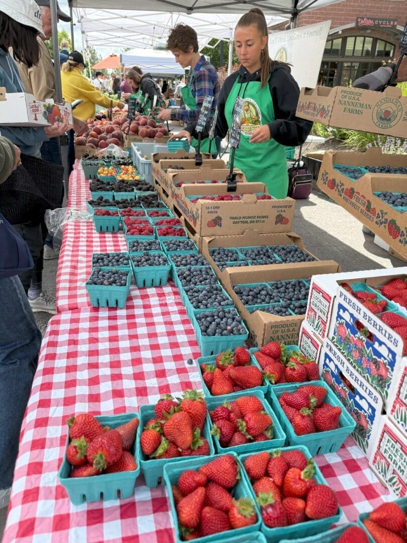 a Farmers Market Stand Displays Baskets of Strawberries Blueberries and Raspberries on a Red and white Checkered Tablecloth Two Vendors in Green Aprons Assist Customers with Stacks of Berry Boxes and Other Shoppers Visible in the Background