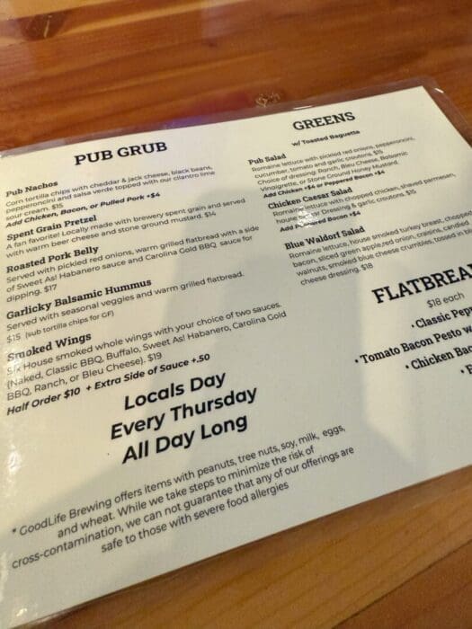 a Restaurant Menu on a Wooden Table Displays Sections Pub Grub Items Like Nachos Hummus Wings Greens Various Salads and Flatbreads Below a Note Mentions Allergen Info and Locals Day Every Thursday All Day Long the Menu Uses Black Text on White Paper