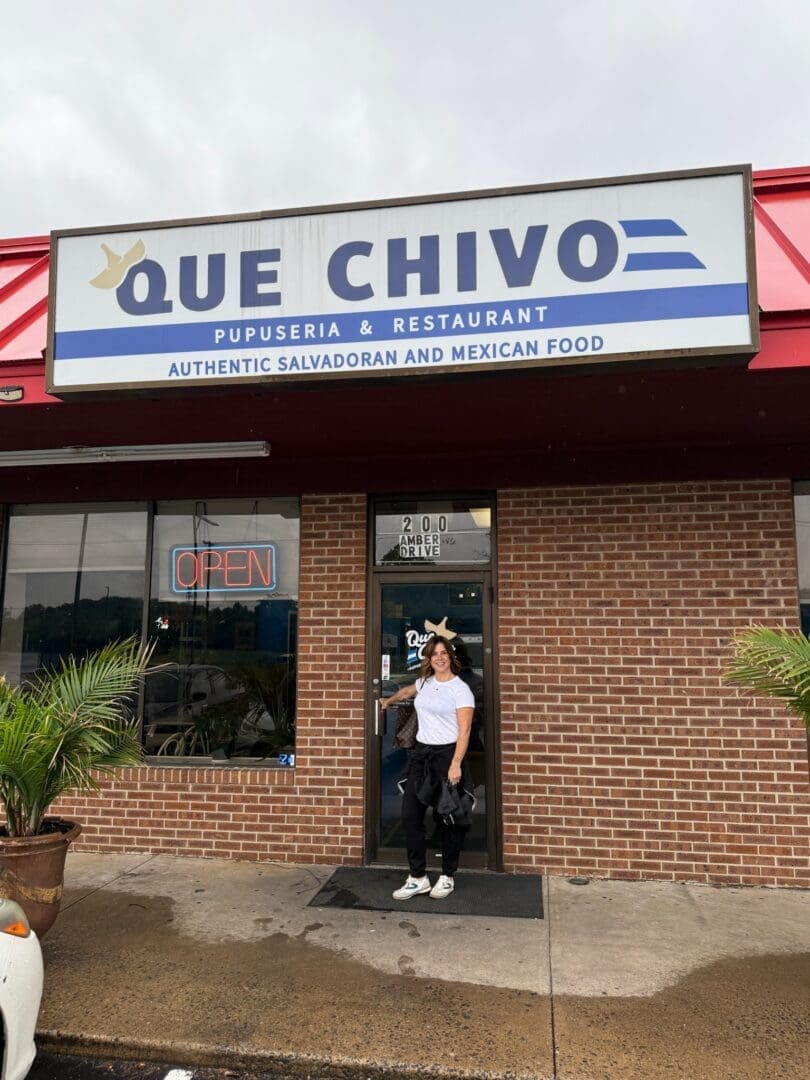 a Woman Stands Smiling in Front of Que Chivo Pupuseria  Restaurant Home to the Best Pupusas in Frederick Md the Sign Above Her Advertises Authentic Salvadoran and Mexican Food Red Trim Brick Walls and Potted Plants Welcome Guests at the Entrance