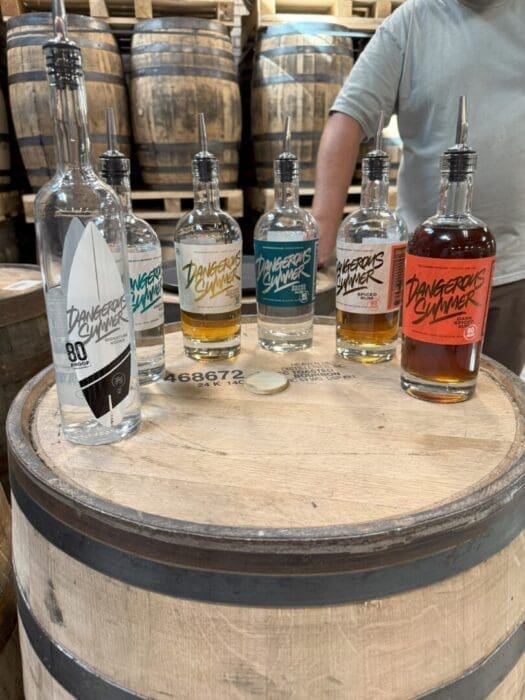 Six bottles of Dangerous Summer spirits, featured at some of the best bars in Frederick, sit on a wooden barrel with vibrant labels. Barrels fill the background while a person in a gray shirt stands behind them, arms partially visible.