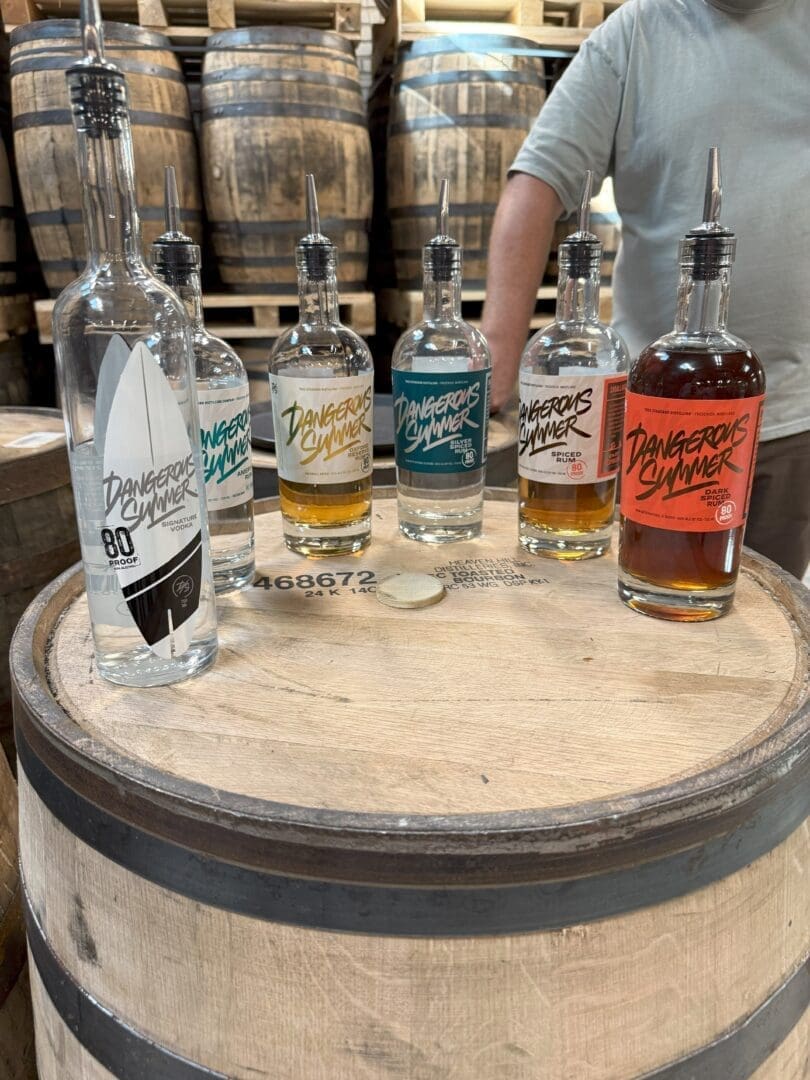 Six Bottles of Dangerous Summer Spirits Featured at Some of the Best Bars in Frederick Sit on a Wooden Barrel with Vibrant Labels Barrels Fill the Background While a Person in a Gray Shirt Stands Behind Them Arms Partially Visible