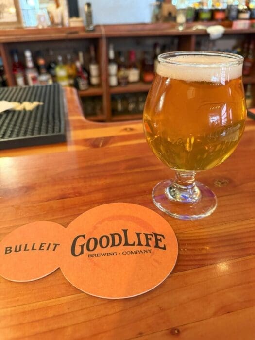 a Round Glass of Golden Beer Sits on a Glossy Wooden Bar Next to an Orange Coaster That Reads Bulleit Goodlife Brewing Company Shelves with Liquor Bottles Are Blurred in the Background the Bar Has a Warm Inviting Atmosphere