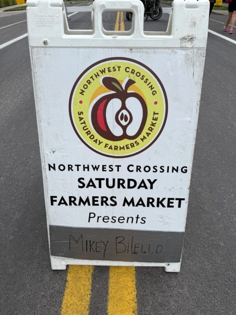 a White Sandwich Board Sign on a Paved Street Displays the Northwest Crossing Saturday Farmers Market Logo with Text Announcing Saturday Farmers Market Presents Mikey Bilello Handwritten at the Bottom a Small Part of a Bicycle is Visible Behind the Sign