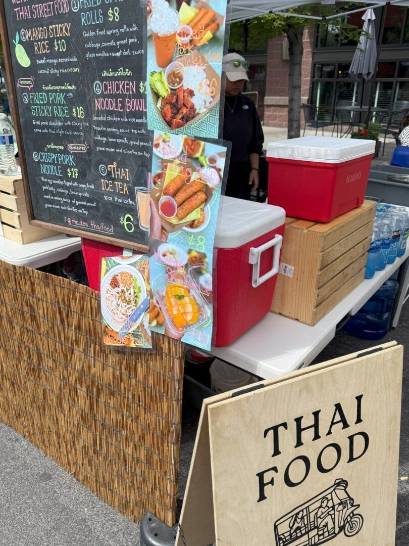 Outdoor Thai Food Stall with a Chalkboard Menu Listing Items Like Mango Sticky Rice and Noodle Bowls Red Coolers Crates and a Staff Member in a Mask and Cap Are Visible Colorful Food Photos and a Large Thai Food Sign Decorate the Stall