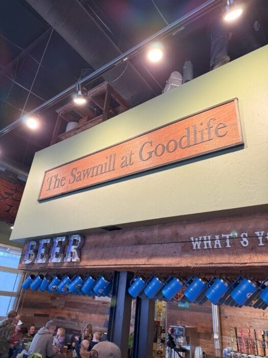 a Wooden Sign Reading the Sawmill at Goodlife is Mounted Above a Bar Area Below a Rustic Beer Sign and Blue Mugs Hang While People Are Gathered and Seated at Tables the Ceiling and Lights Are Visible Above the Green Wall