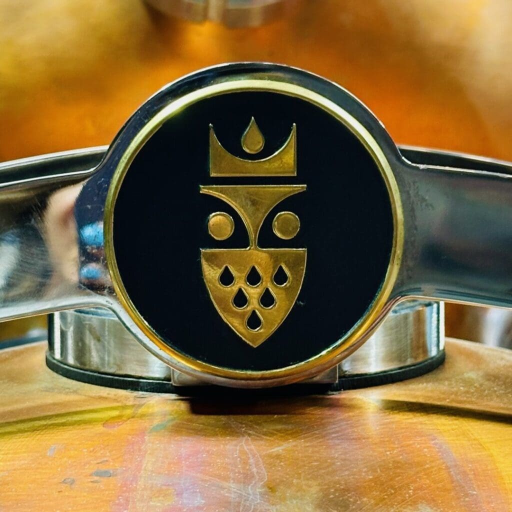 Close Up of a Shiny Metallic Emblem with a Gold Geometric Face and Crown Design on Black Mounted on Copper and Steelits Symmetrical Shapes Echo the Sophistication Found in the Best Bars in Frederick Teardrop Patterns Add Elegance to the Piece