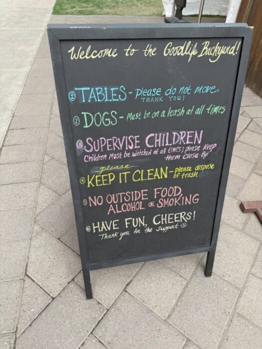 a Black Sandwich Board Sign on a Stone Path Displays Colorful Chalk Writing with Rules Tablesplease Do Not Move Dogsmust Be Leashed Supervise Children Keep It Clean No Outside Food Alcohol or Smoking and Ends with Have Fun Cheers