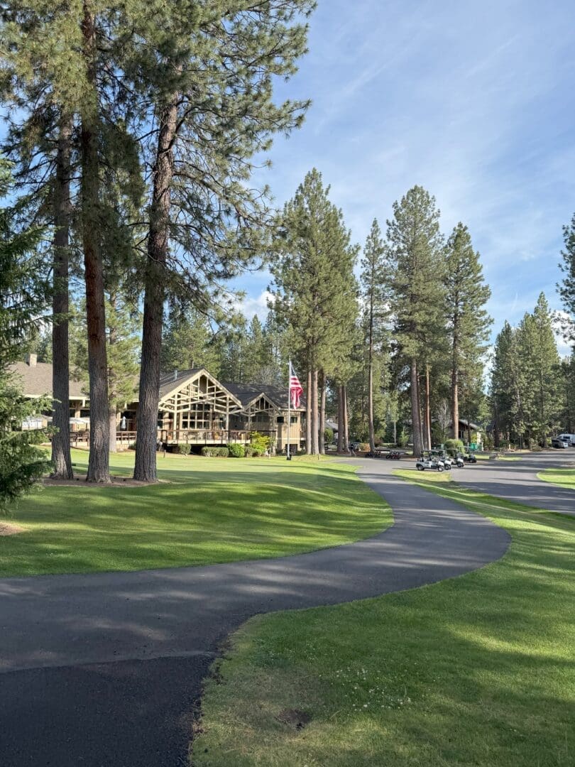 a Winding Paved Path Curves Through a Manicured Lawn with Tall Pine Trees in the Background Theres a Large Rustic Lodge with Many Windows an American Flag on a Pole and Golf Carts Parked Nearby Under a Clear Blue Sky