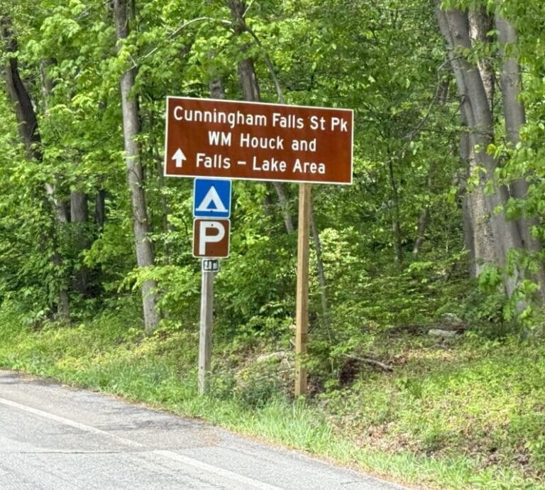 Brown roadside sign reads: Cunningham Falls State Park, WM Houck and Falls – Lake Area with white arrow pointing left. Below are symbols for camping and parking. The sign stands by a paved road amid lush green trees and foliage.