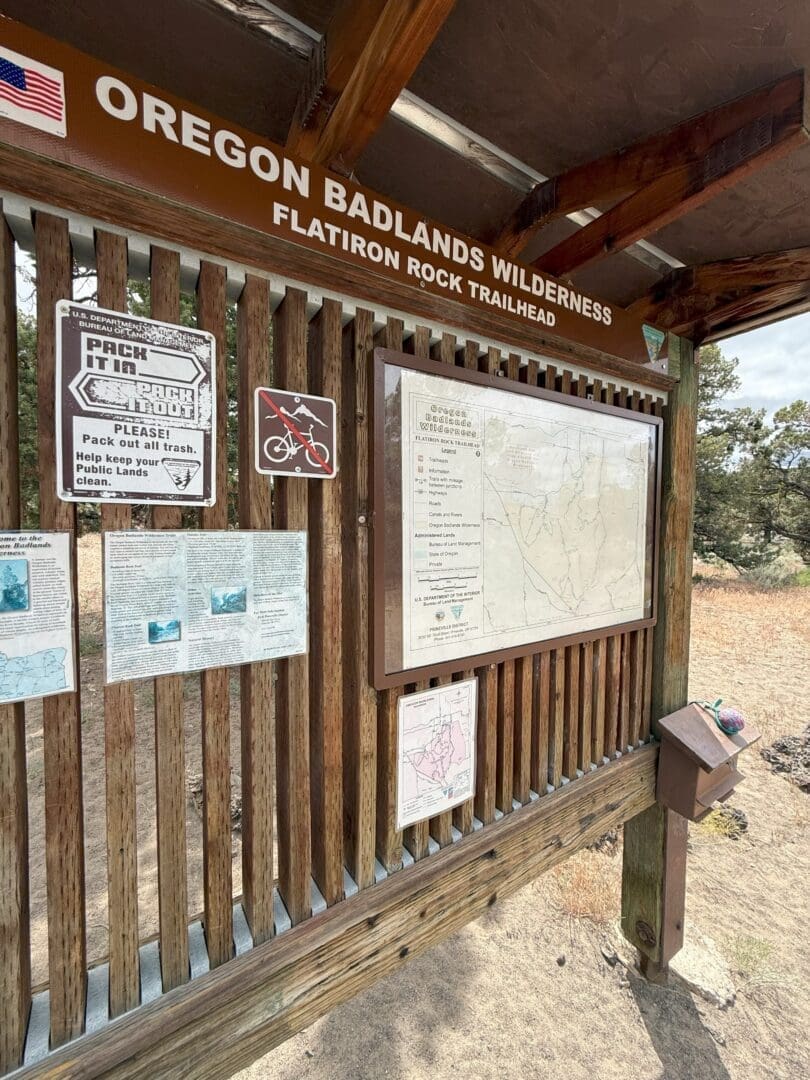 a Wooden Trailhead Sign for Oregon Badlands Wilderness at Flatiron Rock Shows Maps Information Signs and Notices Including a Pack It in Pack It out Reminder Sunlight Shines on the Display Dry Grass and Trees Are Visible in the Background