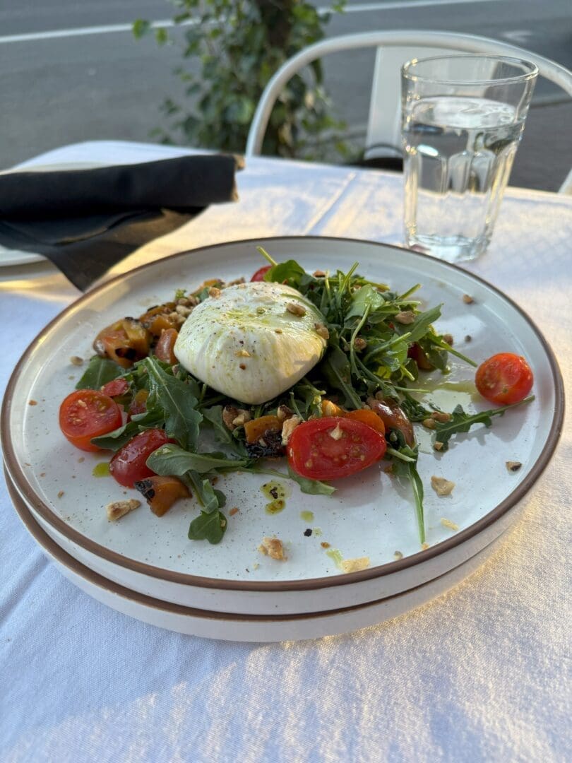 a Plate of Salad with Burrata Cheese on Top Surrounded by Arugula Cherry Tomatoes Nuts and Roasted Vegetables Enjoy This Fresh Dish Outdoors at One of the Top Bend Restaurants with a Glass of Water and a Fork Wrapped in a Black Napkin