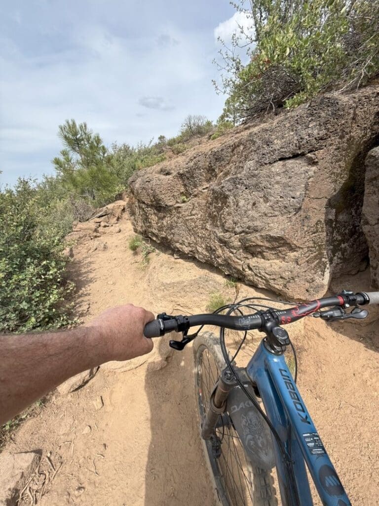 a Person Rides a Blue Mountain Bike on the Narrow Rocky Rimrock Trail Their Left Hand Grips the Handlebar As Rugged Rocks and Green Shrubs Line the Path Under a Partly Cloudy Sky Evoking Outdoor Adventure and Mountain Biking