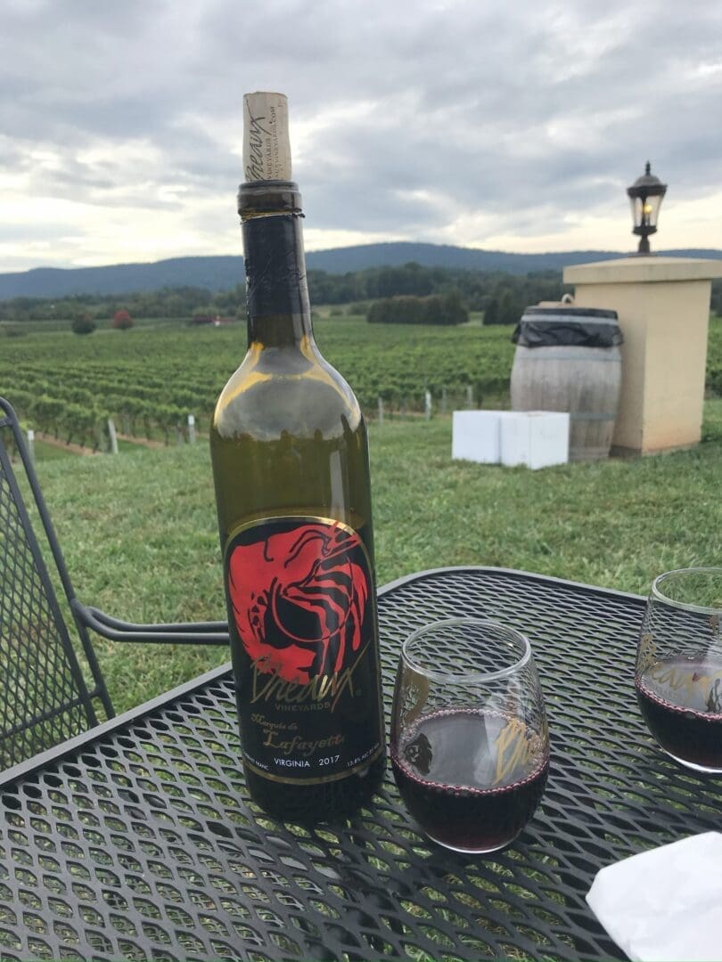 a Bottle of Red Wine Labeled with a Red Scorpion Graphic Stands on a Metal Patio Table at Market Table Bistro Alongside Two Glasses of Red Wine Behind a Vineyard Stretches out Under Cloudy Skies with Green Hills in the Distance and a Barrel and Lantern Nearby