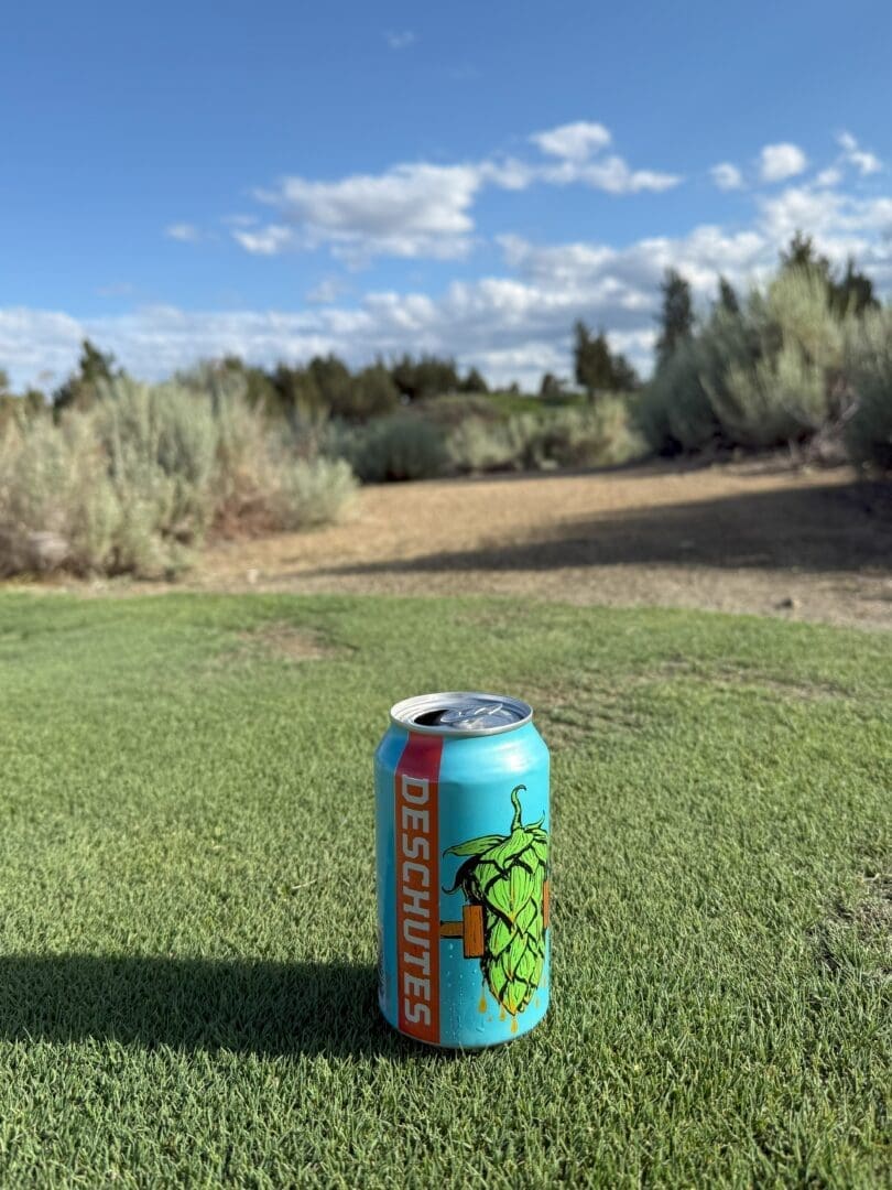 a Blue Deschutes Beer Can with a Green Hop Illustration Stands on Green Grass in the Foreground Reminiscent of a Bend Oregon Golf Course Behind It Shrubs and Dirt Appear Beneath a Bright Blue Sky with Scattered White Clouds