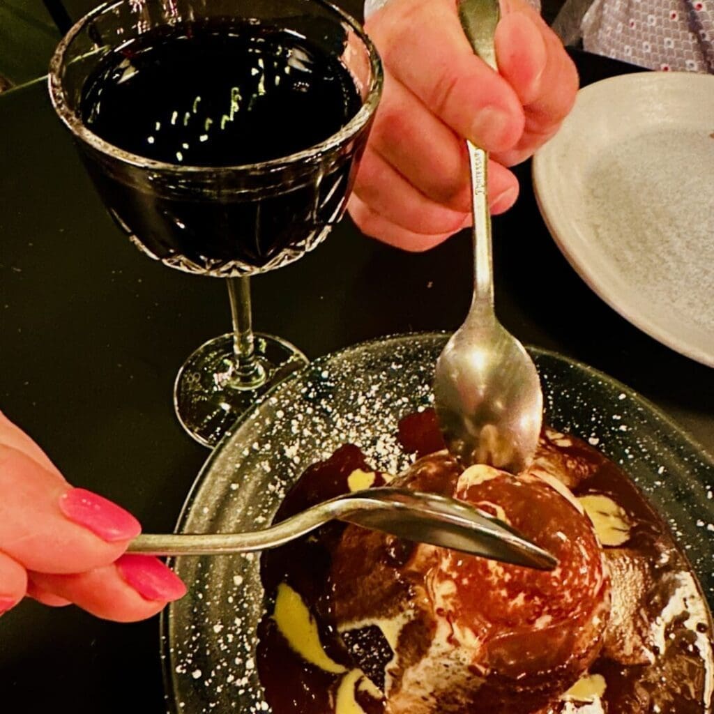 a Close Up of Two People Sharing a Chocolate Dessert with Spoons Next to a Crystal Glass of Red Wine on a Dark Table the Dessert is Topped with Chocolate Sauce and Powdered Sugar One Person Has Pink Nails Both Plates and Hands Are Partially Visible