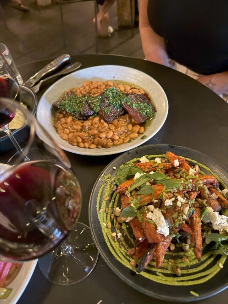 Two Glasses of Red Wine on a Dark Table Beside Two Dishes One with Braised Meat and Beans in a Green Herb Sauce and Another with Roasted Carrots Topped with Greens Nuts and Crumbled Cheese a Person in a Sleeveless Top Sits in the Background