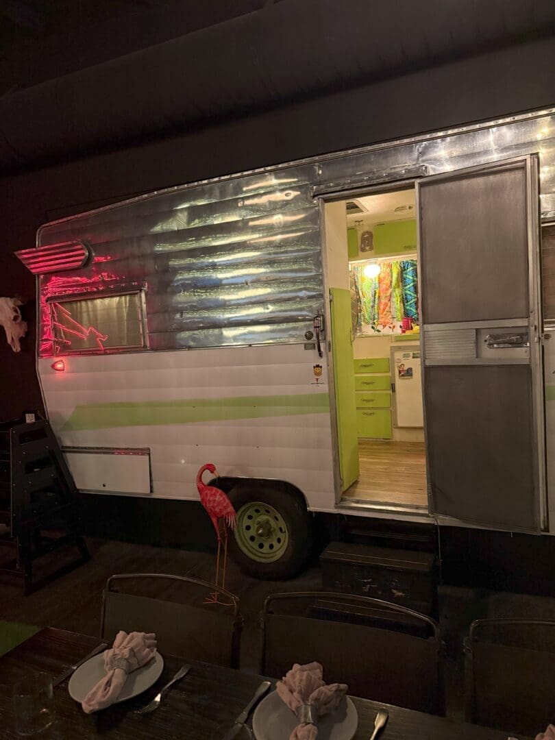a Shiny Silver and White Vintage Camper with a Green Stripe is Parked Indoors Its Door is Open Revealing a Brightly Lit Interior with Colorful Decor a Pink Flamingo Lawn Ornament Stands Outside a Dining Table with Napkins Folded on Plates is in the Foreground