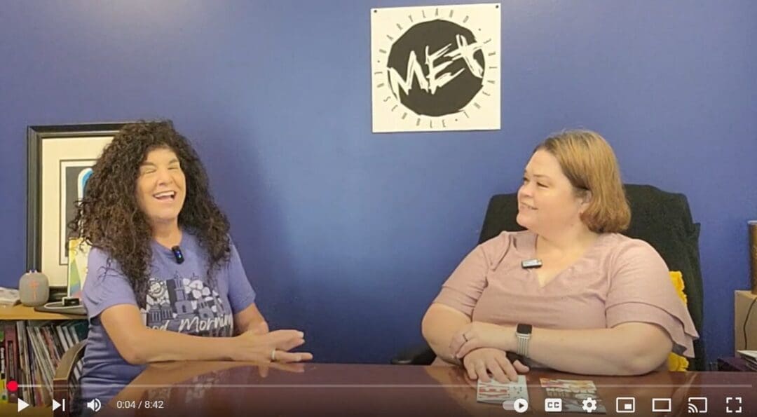 Two women sit at a table against a blue wall, talking and smiling during an Everything Frederick LIVE segment. The woman on the left has long curly dark hair and wears a purple T-shirt; the woman on the right wears a pink top. A logo hangs behind them.