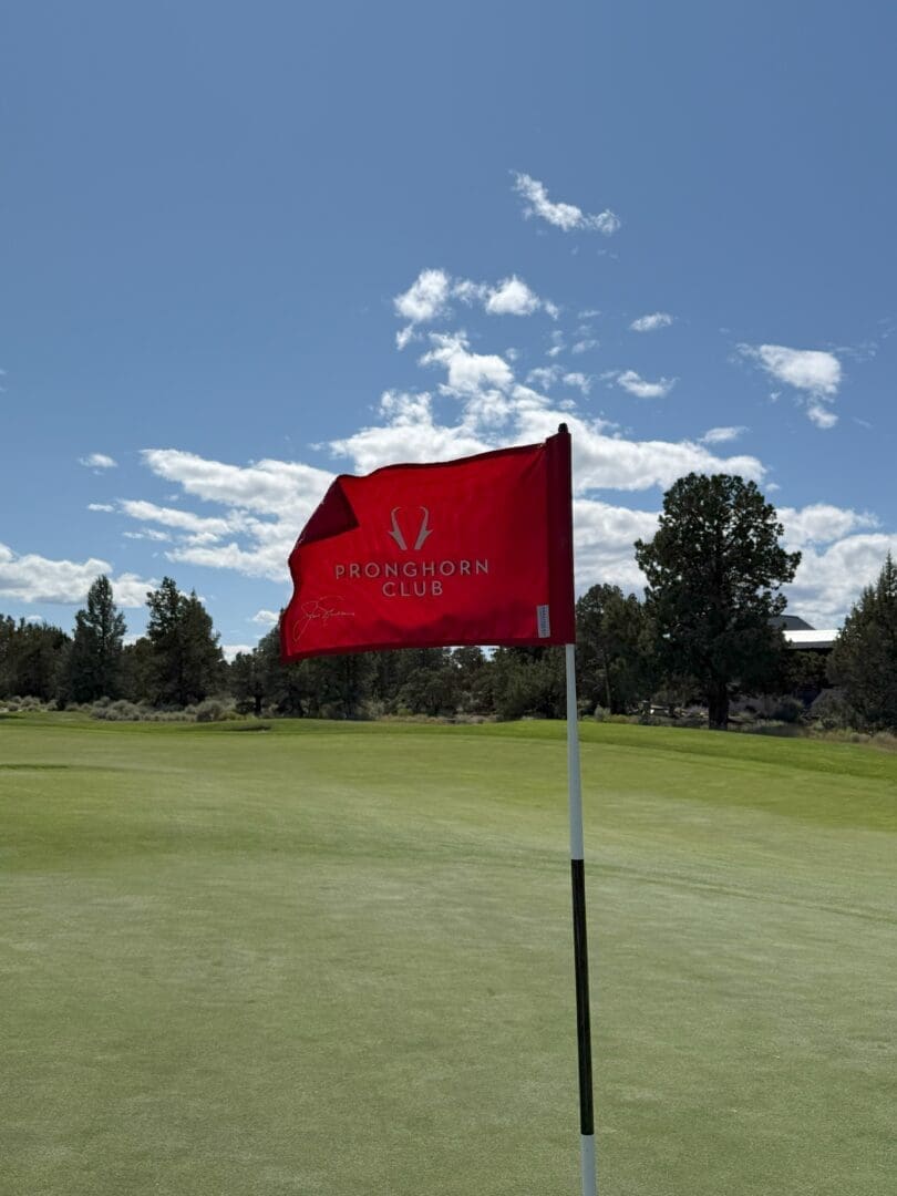 a Red Flag Reading Pronghorn Club Flutters Atop a Flagstick on the Green at Pronghorn Golf Course the Mostly Clear Sky and Tree Lined Background Are Bathed in Bright Sunlight Casting Sharp Shadows Across the Well Manicured Grass