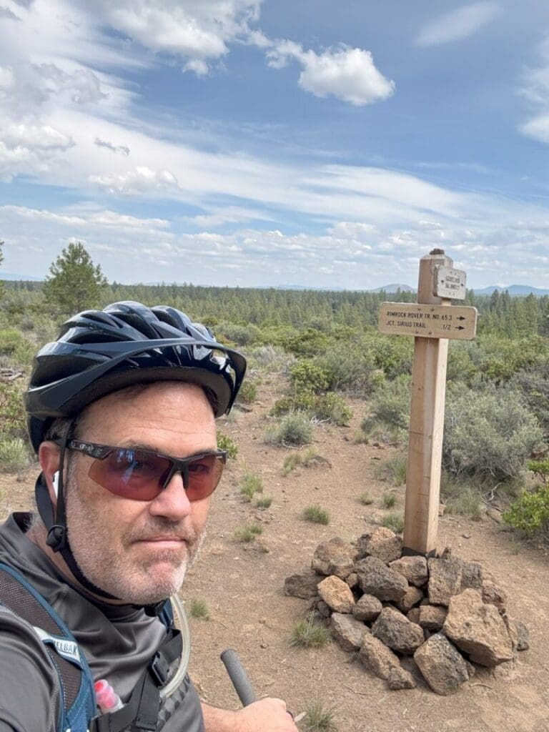 a Man Wearing a Bike Helmet Sunglasses and a Hydration Pack Stands in Front of a Rimrock Trail Sign and a Pile of Rocks on a Dirt Path with Shrubs and Pine Trees Behind Him Under a Partly Cloudy Sky