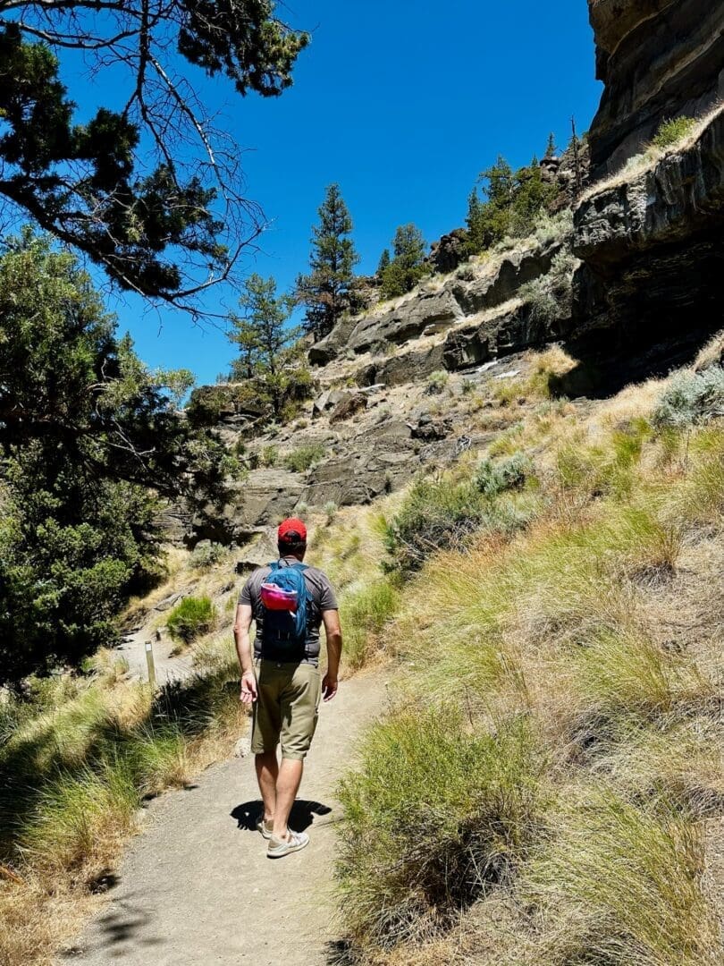 a Person in a Red Cap Blue Shirt Khaki Shorts and a Backpack Hikes Up a Sunlit Dirt Trail Through Dry Grass and Shrubs Near Steelhead Falls Beside Rugged Cliffs and Pine Trees Under a Clear Blue Sky