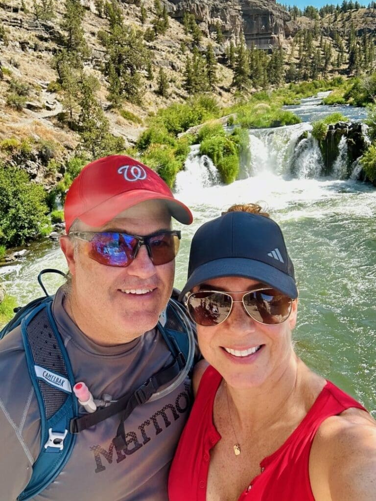 a Smiling Man and Woman Pose for a Selfie Outdoors by Steelhead Falls Near a River and Small Waterfall They Wear Sunglasses Hats and Athletic Clothing Behind Them is Lush Greenery Trees Rocky Terrain and Flowing Water Under a Sunny Sky