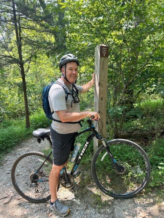 a Man in a Helmet and Backpack Stands Beside a Trek Mountain Bike on a Dirt Trail in the Mt Airy Md Forest He Smiles at the Camera While Holding a Wooden Trail Marker As Sunlight Highlights the Green Foliage Around Him