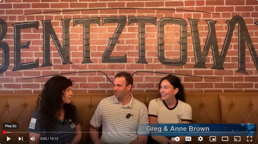 Three people sit on a tan tufted bench in front of a red brick wall with “BENTZTOWN” painted in black letters. Smiling, they appear on Everything Frederick LIVE, with blue lower-third text reading, “Greg & Anne Brown.”.