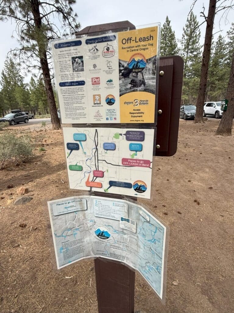a Wooden Signboard in a Forested Parking Area Displays Three Maps and Informational Posters About off Leash Dog Recreation and the Rimrock Trail in Central Oregon with Tall Pine Trees Parked Cars and Dry Brown Soil in the Background
