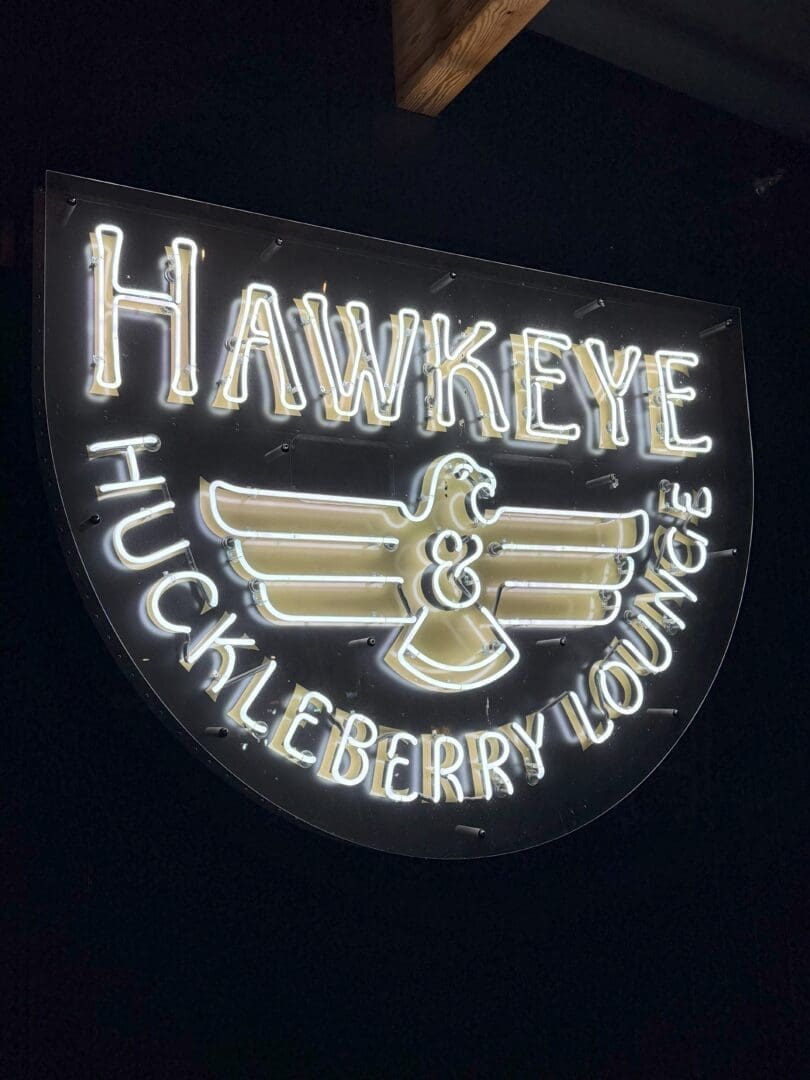 a Glowing Neon Sign Reads Hawkeye  Huckleberry Lounge in White and Gold with a Stylized Bird Spreading Its Wings Set Against a Black Background This Shield Shaped Sign Shines Among Bend Restaurants Outlined in Radiant White Neon