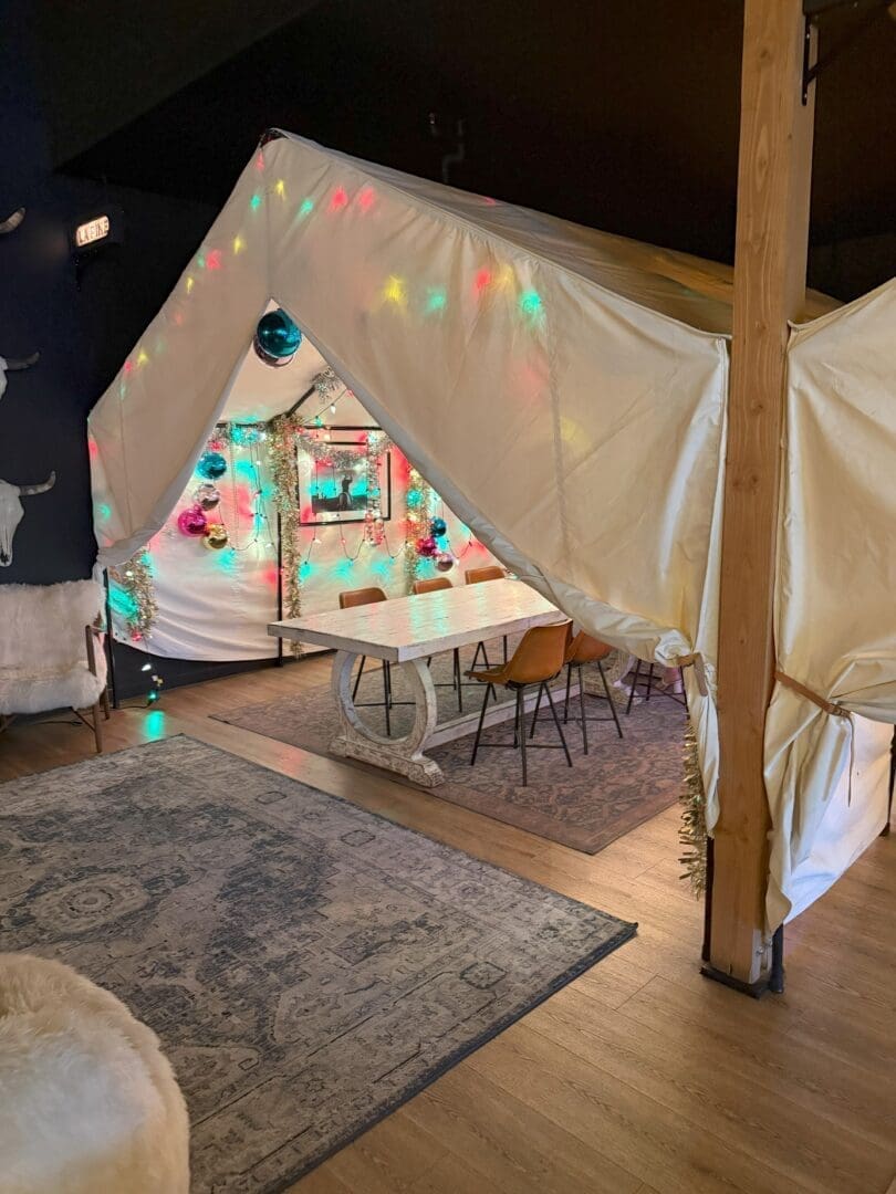 a Cozy Indoor Tent with White Fabric is Decorated with Colorful String Lights and Ornaments Inside a Long Table with Mismatched Chairs Sits on a Rug the Space Has Wood Floors a Carpet and Soft Ambient Lighting Creating a Festive Inviting Atmosphere