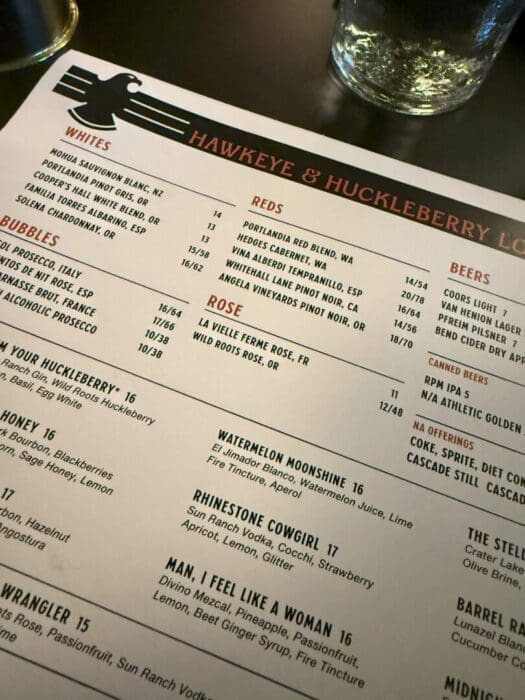 a Close up of a Hawkeye  Huckleberry Lounge Menu on a Dark Table Sections for Wines Beer and Cocktails Are Visible with a Large Black Eagle Graphic at the Top a Glass of Water Sits at the Upper Right Corner Partially Visible