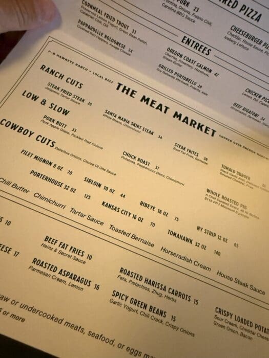 a Close up of a Restaurant Menu Titled the Meat Market Lists Various Steak and Meat Options Including Ranch Cuts Cowboy Cuts Low  Slow and Steak Frites Each Item Has a Description and Price the Menu is Viewed at an Angle with Yellowish Lighting