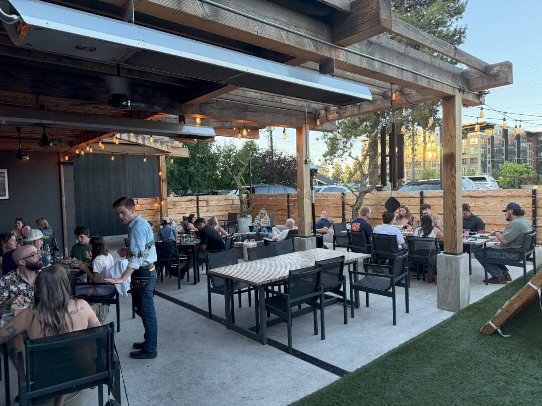 Outdoor Restaurant Patio with People Dining at Tables Under a Wooden Pergola String Lights Hang Above Some Patrons Are Engaged in Conversation a Waiter is Serving Food and Cars Are Visible Beyond the Wooden Fence Its Early Evening with Clear Skies