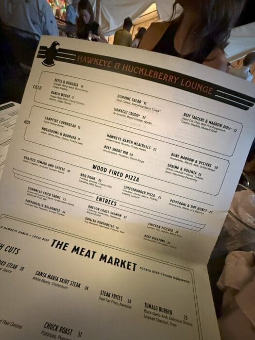 a Person Holds a Large Restaurant Menu at a Dimly Lit Table the Menu Titled Hawkeye  Huckleberry Lounge Lists Starters Hot Dishes Wood fired Pizza Entrees and Meats with Blurred Diners Visible in the Background