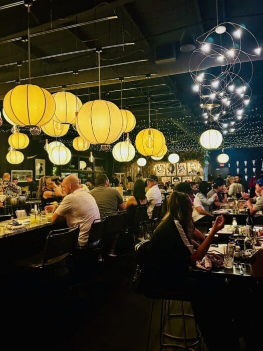 A lively Bend Restaurants bar scene with people seated at tables and the counter, warmly lit by large yellow globe lanterns and string lights overhead. The background features framed art on dark walls and bartenders serving drinks.