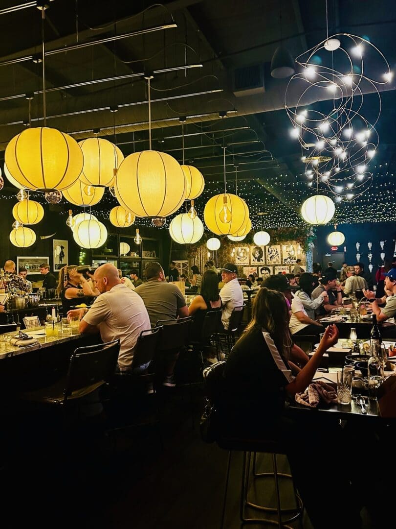 A lively Bend Restaurants bar scene with people seated at tables and the counter, warmly lit by large yellow globe lanterns and string lights overhead. The background features framed art on dark walls and bartenders serving drinks.