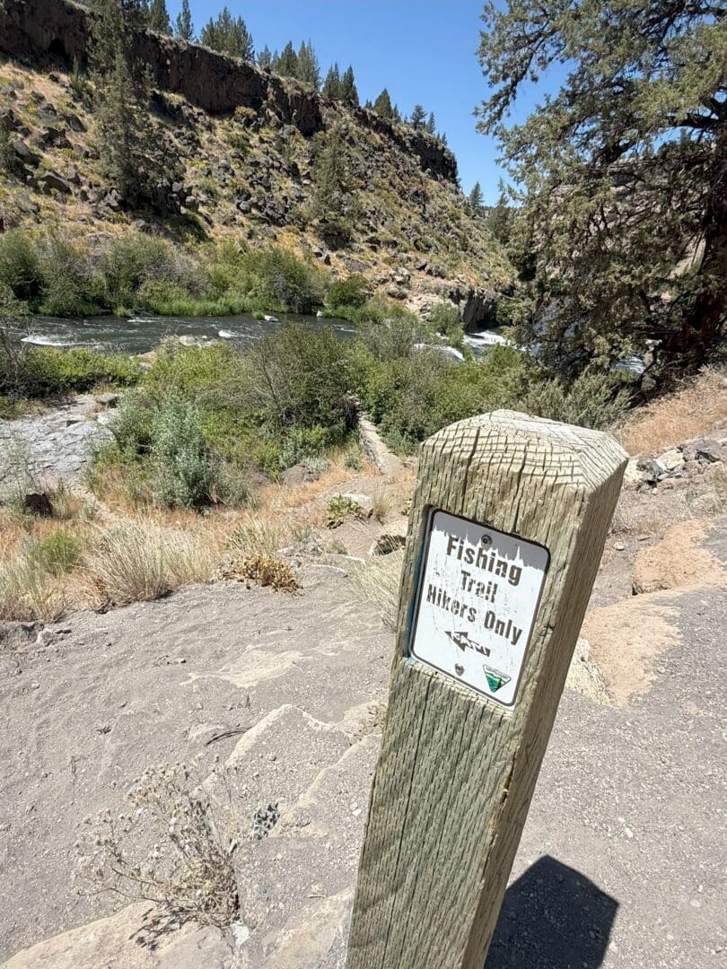 a Weathered Wooden Post with a Fishing Trail Hikers Only Sign Stands on a Dirt Path Overlooking Steelhead Falls Surrounded by Bushes Rocky Terrain and Tall Pine Trees Under a Bright Clear Blue Sky