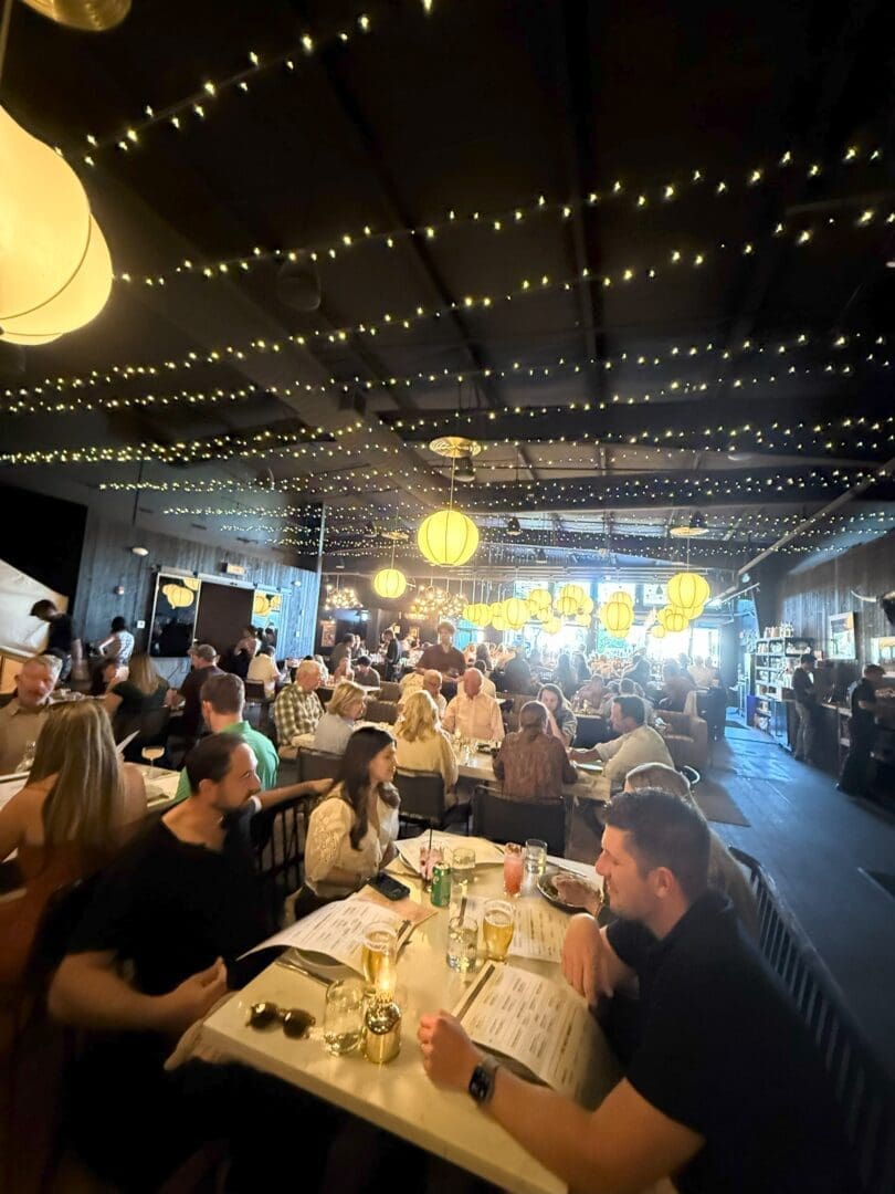 a Lively Restaurant Scene with Many People Dining at Tables Under Warm Hanging Lights and String Lights on the Ceiling the Room is Bustling with Conversation and Menus and Drinks Are Visible on the Tables the Atmosphere Appears Cozy and Inviting