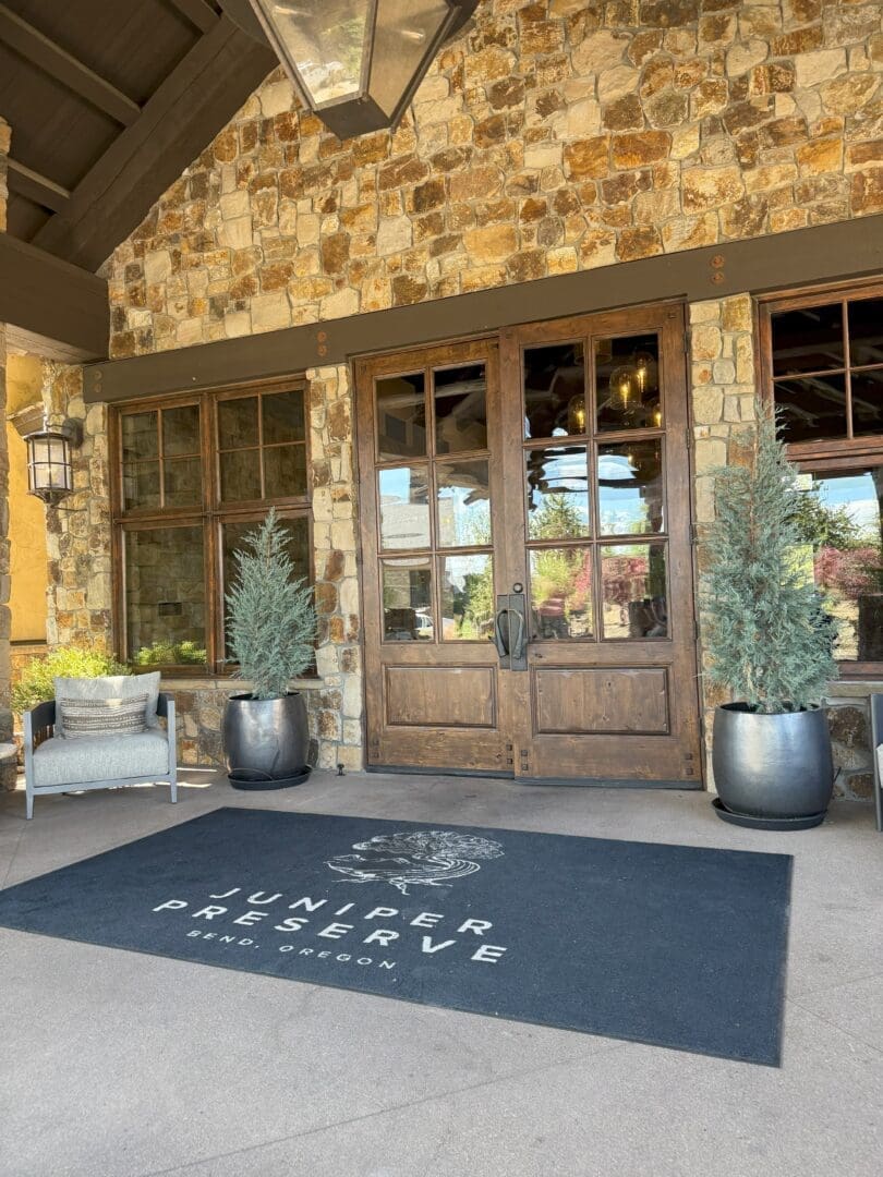 a Stone Building Entrance with Double Wooden Doors Flanked by Large Potted Evergreens Welcomes Visitors to Pronghorn Golf Course a Black Mat in Front Reads Juniper Preserve Bend Oregon Nearby Sits a Grey Cushioned Chair and Lantern Style Wall Light