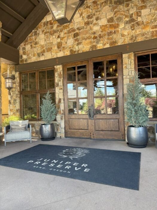 a Stone Building with Large Wooden Double Doors Flanked by Tall Potted Evergreen Trees Welcomes Guests Seeking Where to Stay in Bend Oregon a Black Rug Reads Juniper Preserve Bend Oregon Nearby a Cushioned Chair and Side Table Sit Beneath a Wall Lantern