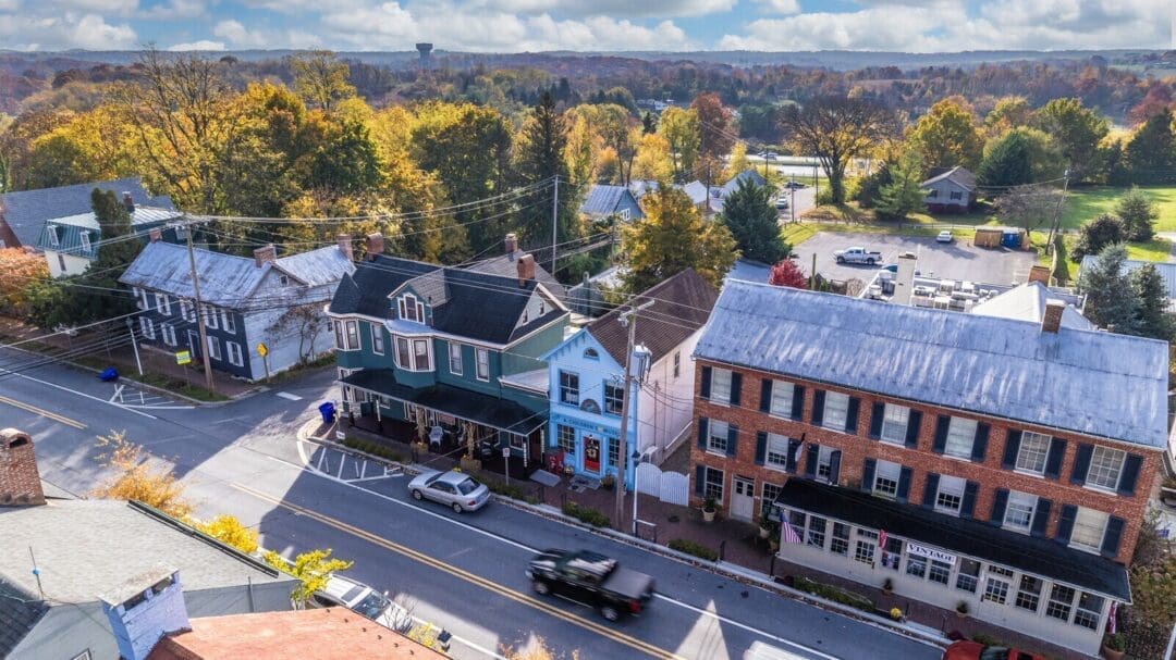 Aerial view of a small town street lined with historic buildings, some brick and some painted wood, surrounded by autumn trees. Cars are parked along the street; a black truck passes by. In the distance, open fields stretch toward hotels in Frederick, MD.