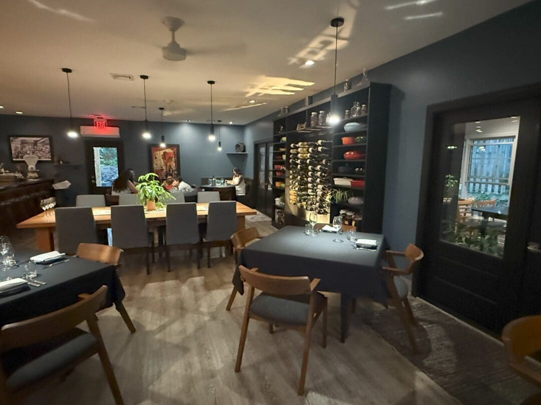 a Modern Restaurant Interior with Dark Walls Wooden Floors and Neatly Set Tables Market Table Bistro Features Wine Racks Shelves with Dishes Contemporary Chairs Soft Lighting and Large Windows Some Tables Are Pushed Together for Group Seating
